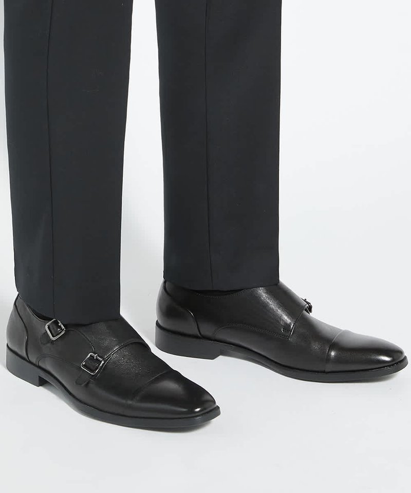 Chaussures habillées en cuir noir à double boucle portées avec un pantalon formel noir sur un fond clair uni.