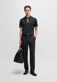 Granatowa koszulka polo z kontrastowym paskowym kołnierzykiem i mankietami, noszona z dopasowanymi czarnymi spodniami i błyszczącymi czarnymi butami. Czarna skórzana torba podróżna.