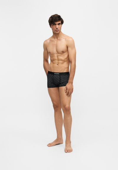 Jeune homme aux cheveux foncés debout, pieds nus, portant un boxer noir à motifs sur un fond clair uni.