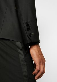 Schwarzer maßgeschneiderter Blazer mit glatter Textur, ausgestattet mit großen schwarzen Knöpfen und kontrastierenden grünen Nähten an den Manschetten. Schwarze Hosen sind teilweise sichtbar.