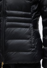 Veste matelassée noire avec couture horizontale, fermeture éclair frontale et poche latérale, visible de la taille jusqu'au milieu de la cuisse.