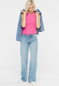 Rosa ärmelloses Oberteil kombiniert mit hellblauen weit geschnittenen Jeans und weißen Sneakers, mit einer Jeansjacke über die Schultern gelegt.