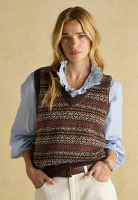 Ženska z dolgimi blond lasmi nosi vzorčasto rjavo pleteno brezrokavnik čez svetlo modro srajco z nabranim ovratnikom ter smetanaste hlače, roke ima v žepih.