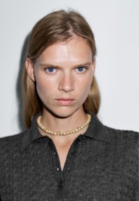 Szara koszula z kołnierzykiem o teksturowanej tkaninie; model nosi jasny naszyjnik z koralików. Tło jest jednolicie białe. Niebieskie oczy są wyraźnie widoczne.