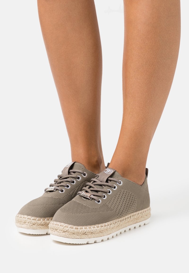 Paire de baskets en maille taupe avec semelles à détails en corde, portées sur des jambes nues sur fond blanc.
