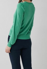 Pull vert en tricot avec manches longues, poignets côtelés et col rond, assorti à un pantalon foncé. Texture lisse, détails subtils aux épaules.
