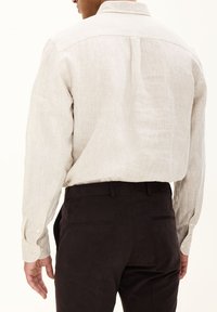 Camicia in lino beige chiaro con maniche lunghe, dettaglio delle pince sul retro e polsini con bottoni, abbinata a pantaloni scuri e aderenti.