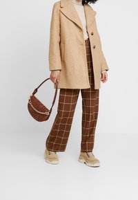 Beige textured coat with large lapels, brown checkered trousers, beige sneakers, and a brown patterned handbag with a chain strap.