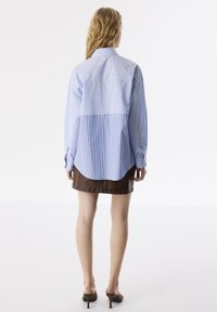 Helleblaue Button-up-Bluse mit vertikalen Streifen, längerer Rückseite und umgeschlagenen Ärmeln. Kombiniert mit einem braunen Leder-Minirock.