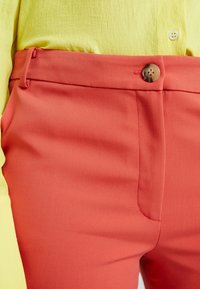 Pantalon corail en tissu texturé avec une finition lisse, doté d'une fermeture à un bouton et de poches latérales, associé à un haut jaune.