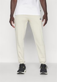 Puma joggebukser i lys beige, med en tætsiddende pasform, elastiske manchetter og et logo på venstre lår. Båret med sorte sportssko.