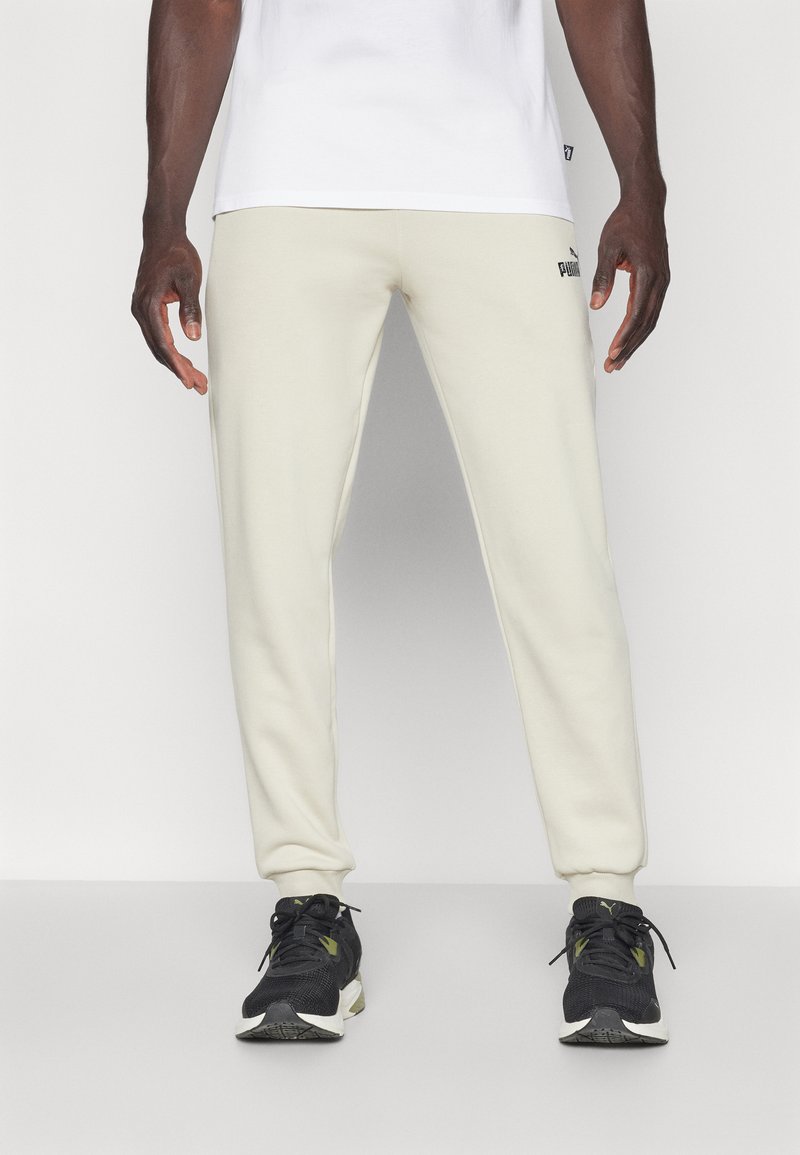 Puma joggebukser i lys beige, med en tætsiddende pasform, elastiske manchetter og et logo på venstre lår. Båret med sorte sportssko.