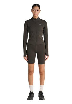 Vrouw staand, gekleed in een bijpassend zwart jack met rits en biker shorts, witte sokken en zwarte sneakers, gericht naar voren op een witte achtergrond.