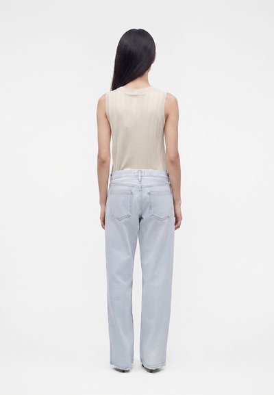 Γυναίκα με μακριά μαύρα μαλλιά στέκεται με την πλάτη, φορώντας ένα μπεζ αμάνικο τοπ και ανοιχτό μπλε φαρδιά τζιν μπροστά σε απλό φόντο.