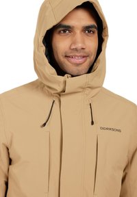 Beige vattentät jacka med huva, utrustad med dragkedja framtill, elastiska snoddar och en subtil logotyp på bröstet. Slät syntetisk tyg.