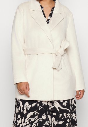 Femme portant un manteau beige ceinturé par-dessus une robe fleurie noire et blanche, debout, les mains détendues le long du corps.