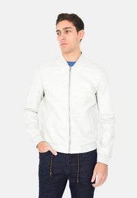 Giacca bomber in pelle bianca con chiusura a zip, colletto e polsini a coste, due tasche laterali, indossata sopra una camicia blu, abbinata a jeans scuri.