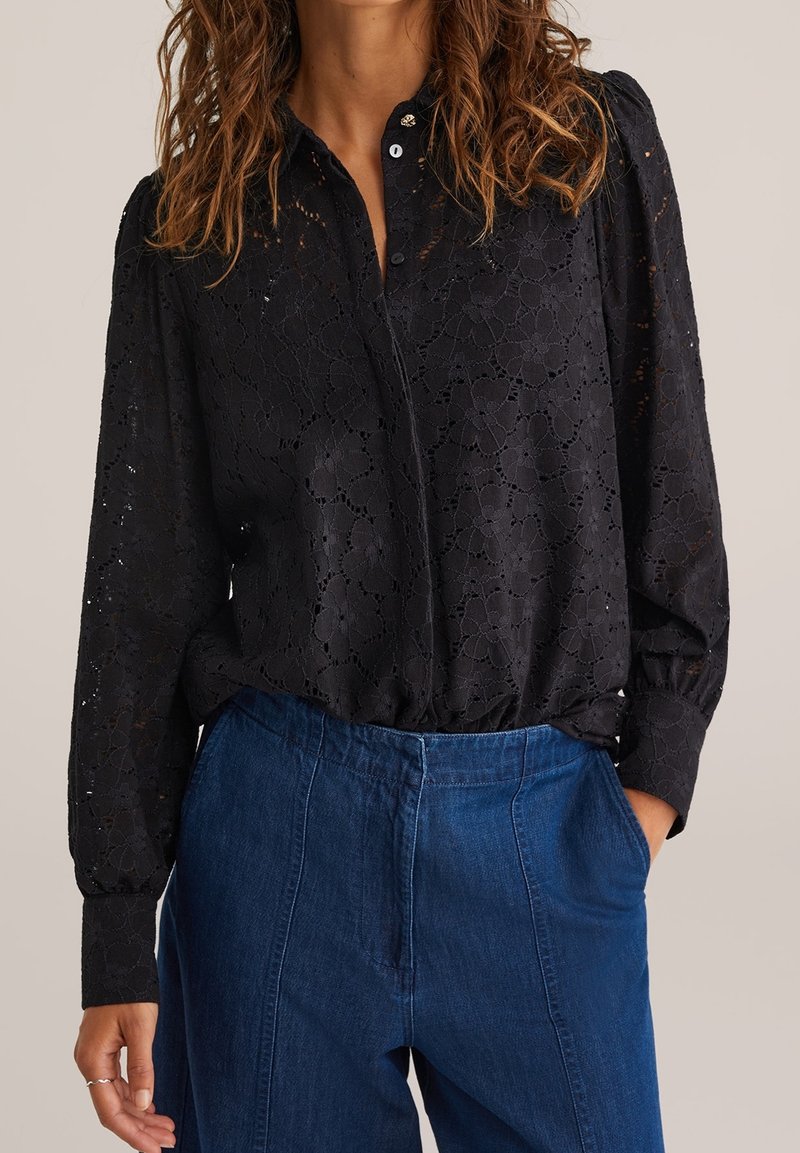 Femme portant un chemisier en dentelle noire à manches longues avec motifs floraux et un pantalon en jean bleu taille haute, une main dans la poche.