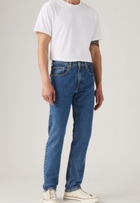 Homme portant un jean droit bleu, un t-shirt blanc à col rond, des baskets blanches et un tatouage noir partiel sur l'avant-bras gauche sur un fond uni.