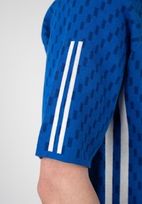 Camiseta de manga corta de punto azul con patrón rectangular azul oscuro y dos rayas verticales blancas en el brazo sobre un codo doblado.