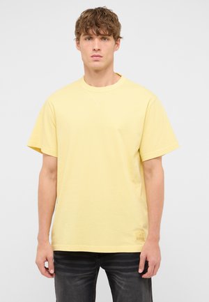 Young man wearing a plain light yellow short-sleeve t-shirt and dark jeans standing against a neutral background.