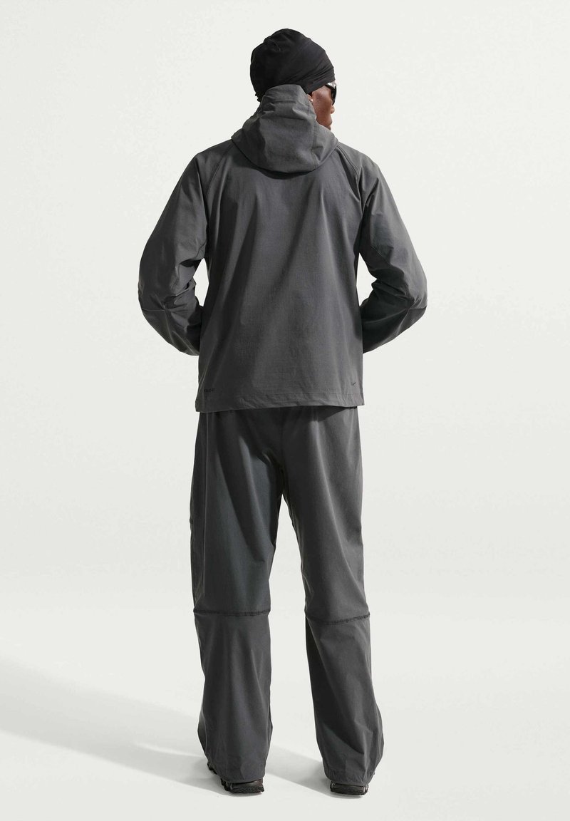 Man facing away wearing dark gray hooded jacket, matching pants, black head covering, and sunglasses against plain light background.