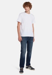 T-shirt bianco a maniche corte, abbinato a jeans blu scuro. Il modello indossa scarpe sportive bianche e nere, in piedi con una postura rilassata.