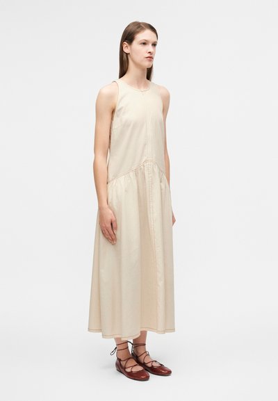 Μπεζ αμάνικο φόρεμα με ραφή στη μέση, κομμένο μέχρι τον αστράγαλο. Συνδυάζεται με καφέ επίπεδα δερμάτινα παπούτσια με κορδόνια. Ομαλή υφή.