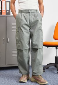 Zielone spodnie cargo wykonane z bawełnianej mieszanki, wyposażone w boczne kieszenie, luźny krój i zapinane na guziki. Połączone z brązowo-różowymi sneakersami.