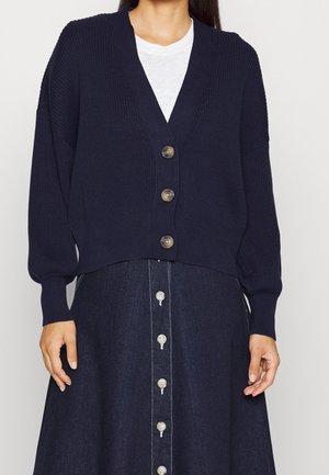 Kvinde iført en mørkeblå knappet cardigan over en hvid top kombineret med et mørkt denim knap-foran nederdel.