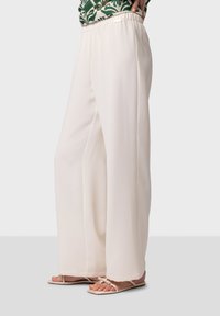 Wijdvallende broek van lichtgewicht, crèmekleurige stof met een elastische tailleband. Gepaarde met eenvoudige, beige sandaaltjes met bandjes.