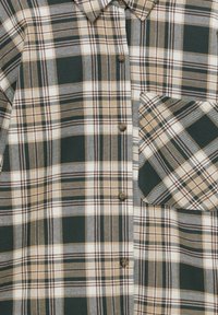 Groen, beige en witte geruite blouse met knopen aan de voorkant en een enkele borstzak op een houten oppervlak.