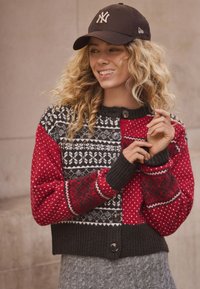 Punainen ja musta neulottu villapaita kuvioiduilla hihoilla, napillinen suljenta ja resorit hihansuissa. Yhdistetty mustaan lippikseen, jossa valkoinen logo.