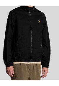 Svart zip-up jacka med ståkrage, elastiska ärmslut, två framfickor och en liten gyllene logotyp på bröstet. Slät tygtextur.