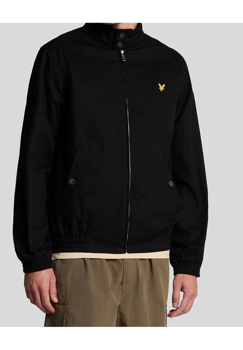 Svart zip-up jacka med ståkrage, elastiska ärmslut, två framfickor och en liten gyllene logotyp på bröstet. Slät tygtextur.