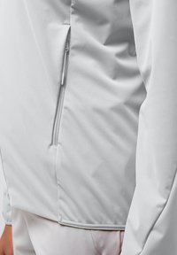 Light gray athletic jacket featuring a side zipper, smooth texture, and sleek design, partially shown from an angled perspective.
