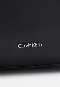 Logo Calvin Klein en lettres métalliques argentées sur une surface lisse en cuir noir, probablement partie d’un sac à main ou d’un accessoire.