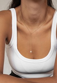 Gouden gedraaide ketting met een parel hanger, gedragen over een wit aansluitend tanktop, rustend op blote huid.