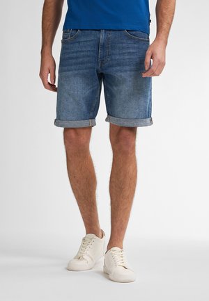 Shorts in denim di blu medio con orlo arrotolato. Indossati con una camicia blu e sneakers beige. Presentano cinque tasche e un leggero sbiadimento.