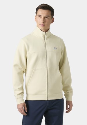 Hombre con chaqueta cremosa con cremallera y cuello alto, y pantalones azul marino, de pie sobre un fondo claro y liso.