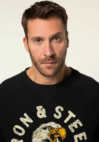 Sudadera negra con cuello redondo, que presenta un gráfico desgastado de un águila y el texto "IRON & STEEL" en amarillo y blanco.