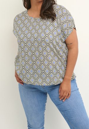 Woman in blue jeans and short-sleeve blouse with black and blue geometric diamond pattern, standing with one hand on hip.