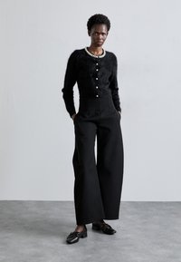 Cárdigan de mohair negro con botones de perlas, combinado con pantalones de tiro alto y pernera ancha en negro y zapatos negros con detalle de lazo.