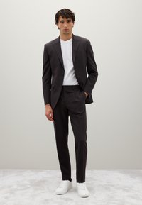 Pak van donkergrijze stof met een getailleerde pasvorm, gecombineerd met een witte T-shirt met ronde hals en witte sneakers. Glad textuur, minimale details.