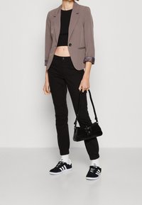 Grå blazer med upprullade ärmar, svart crop top, svarta slim-fit byxor och svarta sneakers med vita ränder. Bär en liten svart handväska.