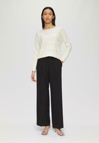 Weißer, wellenförmiger Pullover kombiniert mit weit geschnittenen schwarzen Hosen. Das Modell trägt silberne Absatzsandalen, vor einem neutralen Hintergrund präsentiert.