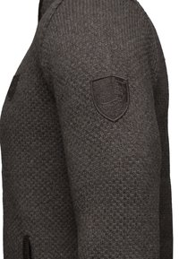 Mörkgrå stickad tröja med ett strukturerat mönster, som har ett broderat emblem på vänster axel och sidofickor för extra detalj.