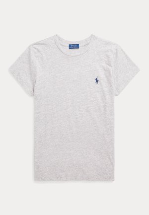 Donna in piedi che indossa una maglietta grigia a maniche corte, jeans blu a gamba larga con cintura e mocassini marroni, sfondo neutro.