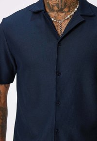 Chemise courte à manches en bleu marine avec des rayures verticales texturées, design à col et fermeture à boutons sur le devant. Collier en chaîne visible comme accessoire.