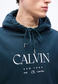 Εγγύς λήψη ενός ατόμου που φοράει σκουρόχρωμο μπλε-πράσινο φούτερ με κουκούλα και λευκή κεντητή στάμπα στο στήθος με το λογότυπο "CALVIN NEW YORK 1968 CK Calvin Klein".